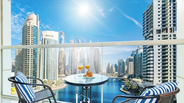 Ein Balkon mit Glastisch, zwei Stühlen und Blick auf eine Skyline mit Hochhäusern und einem Gewässer.