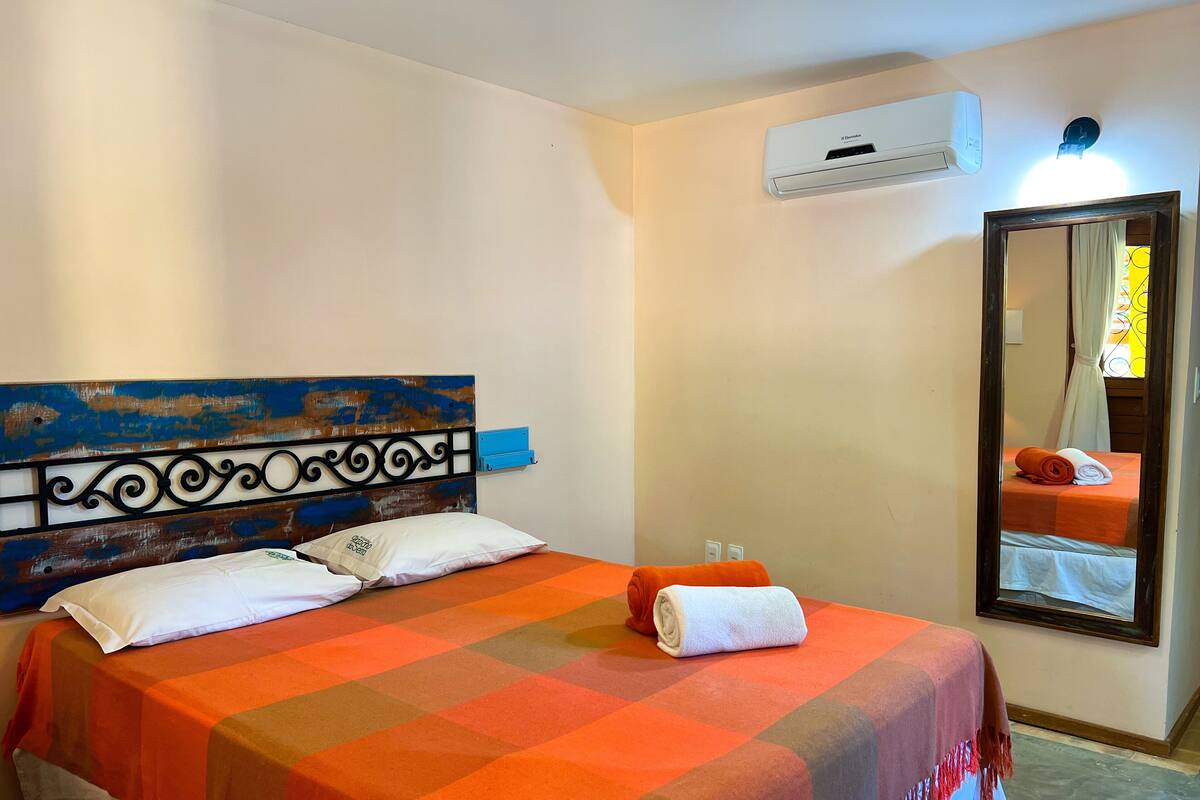 A hotel room with a bed, a headboard with a decorative metal frame, an air conditioning unit, a mirror, and a window with curtains.