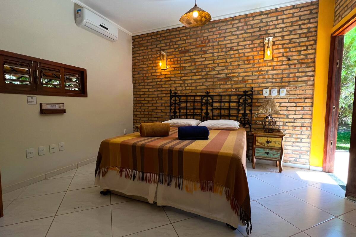 A bedroom with a brick wall, a bed with a striped blanket, a bedside table with a lamp, and a window with a view of greenery.