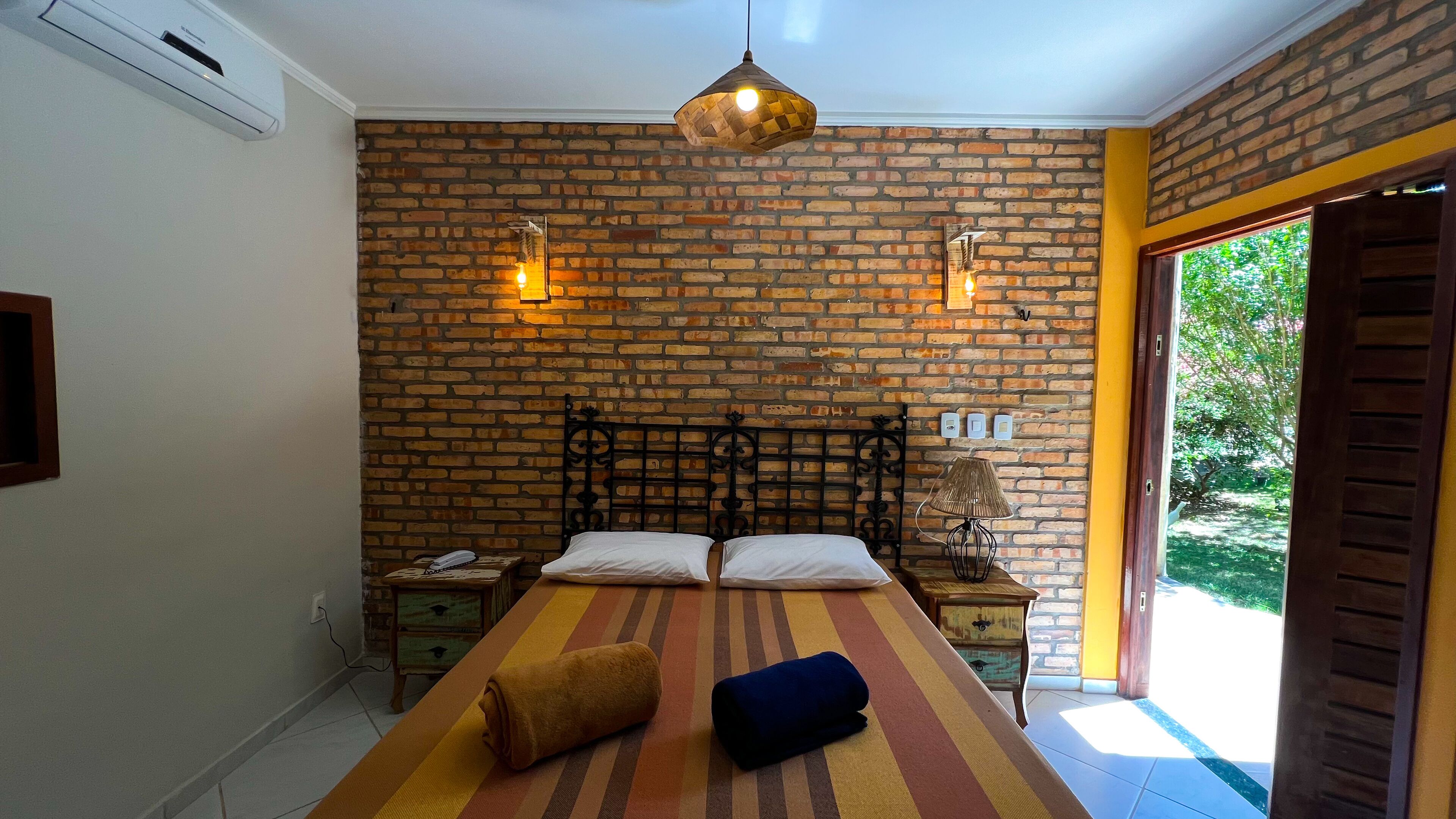 A bedroom with a brick wall, a bed with two pillows, a striped rug, a lamp, and a window with a view of greenery.