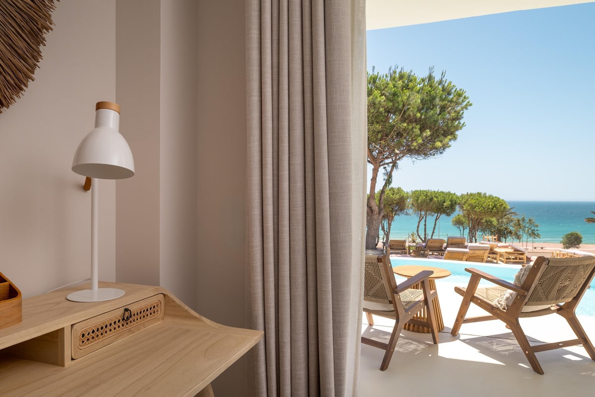 A room with a wooden desk, a white lamp, and a view of a pool and beach.
