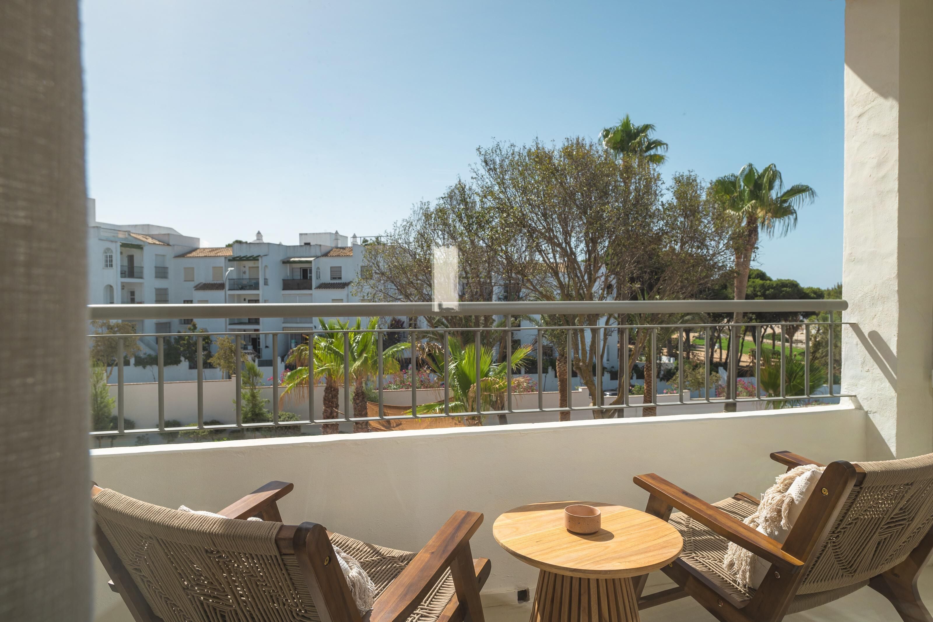Un balcón con mobiliario de mimbre y una pequeña mesa, con vistas a una zona residencial con palmeras y edificios blancos.