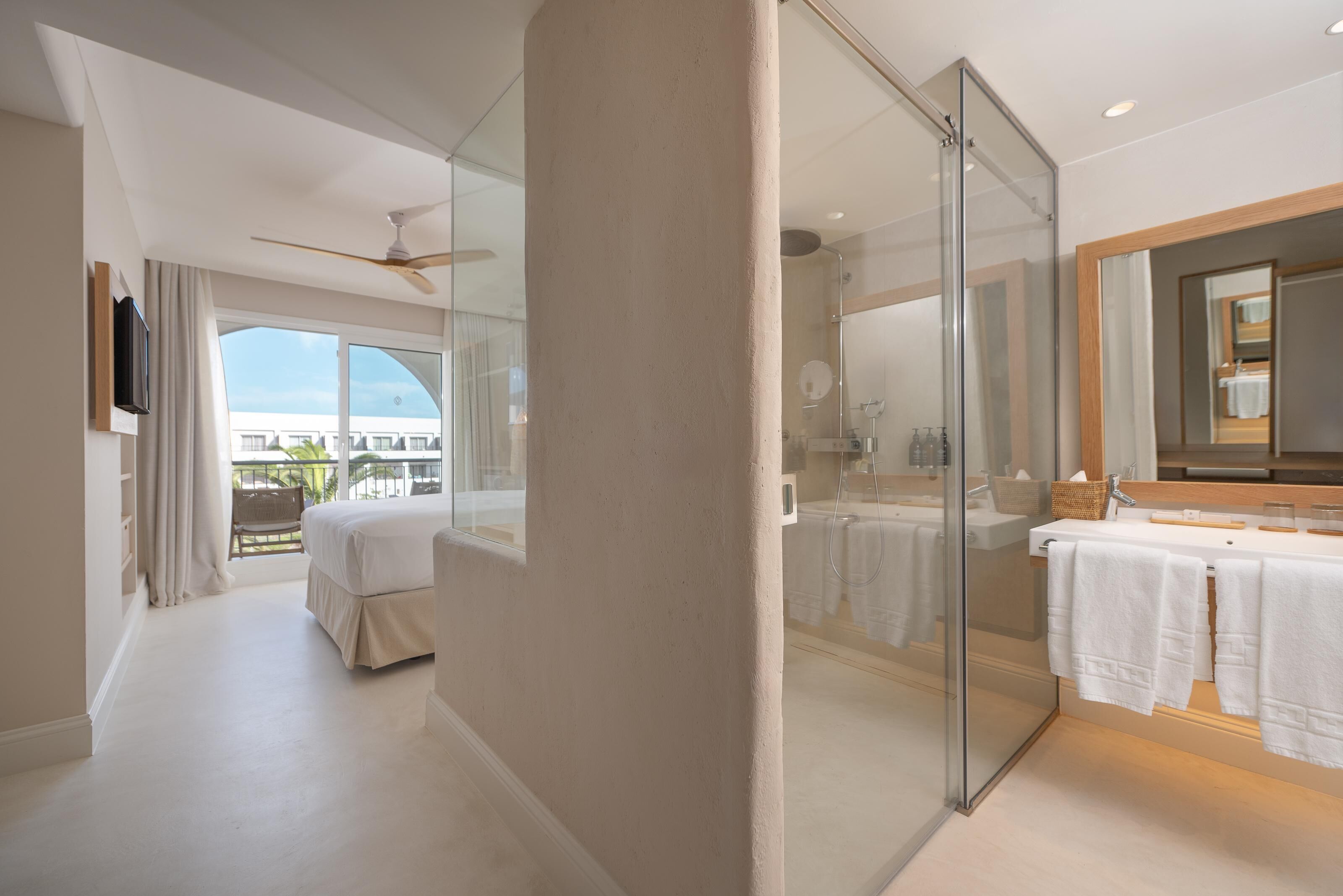 A modern hotel room with a large bed, a glass shower enclosure, a vanity with a mirror, and a balcony with a view.