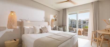 A modern hotel room with a large bed, a ceiling fan, a balcony with a table and chairs, and a view of the city.