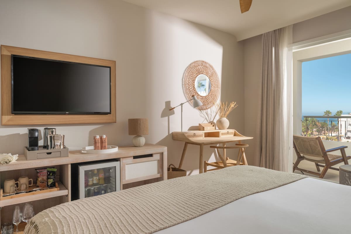 A hotel room with a large bed, a flat-screen TV, a desk with a lamp, a small refrigerator, and a view of the ocean.