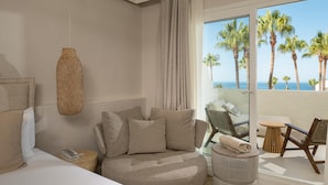Ein Hotelzimmer mit gebogenem Sofa, einem kleinen runden Tisch und einem Balkon mit Blick auf Palmen und das Meer.