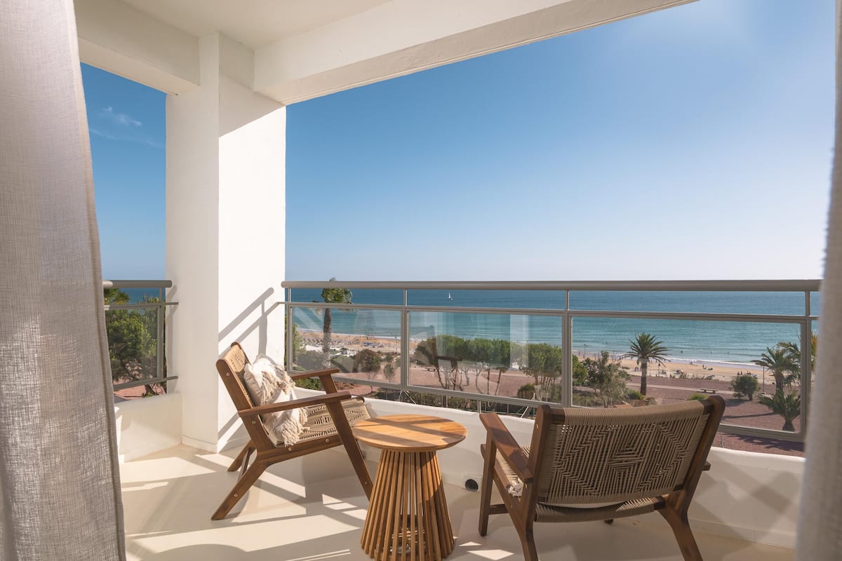 A balcony with a wooden table and two wicker chairs, overlooking a beach with palm trees and a clear blue sky.