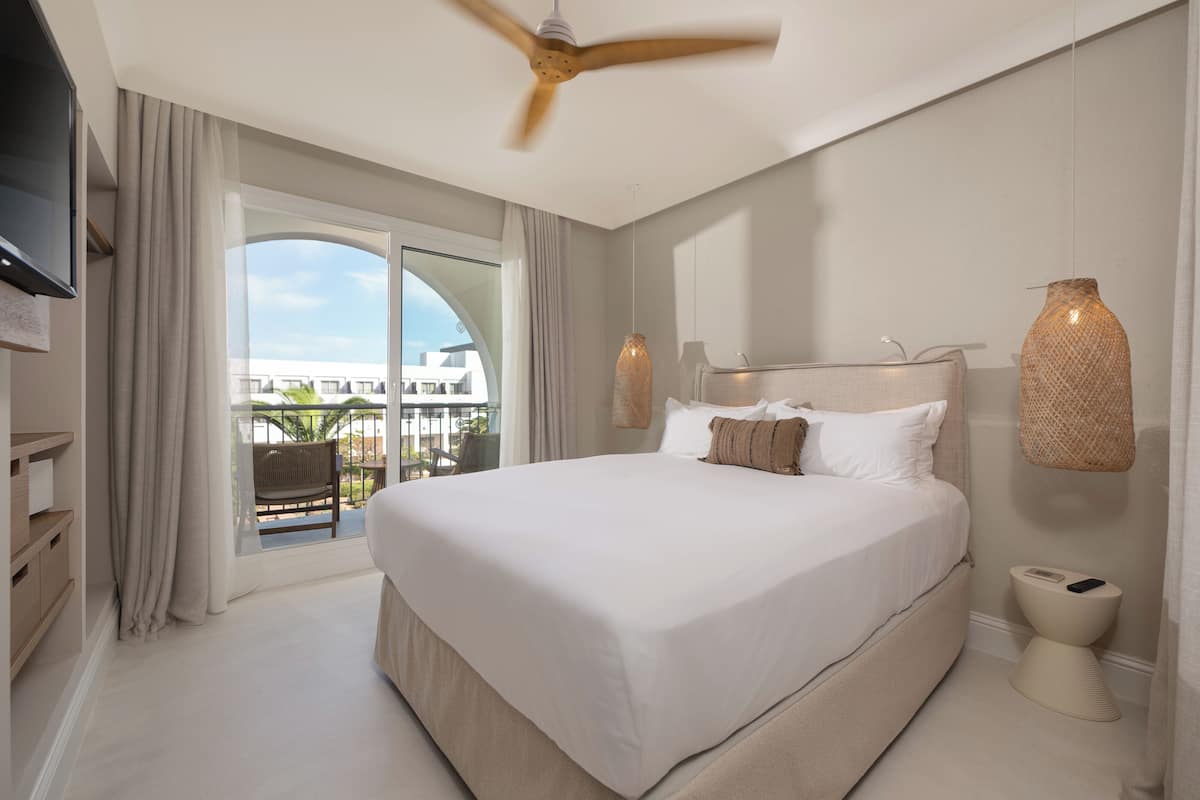 A spacious bedroom with a large bed, a ceiling fan, a TV, and a balcony with a view of buildings.