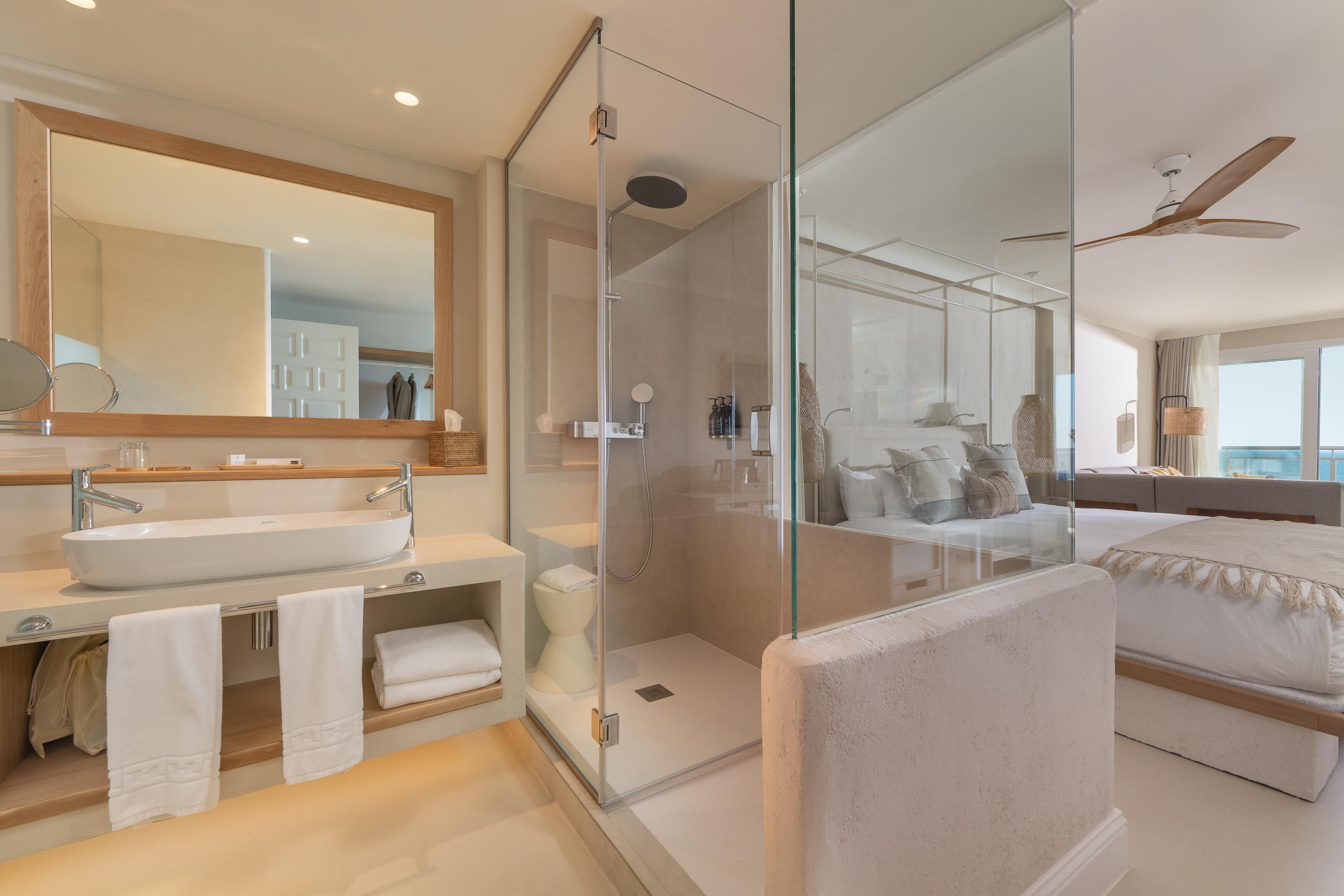 A modern bathroom with a glass-enclosed shower, a large mirror, a white sink, and a bed with a white headboard.