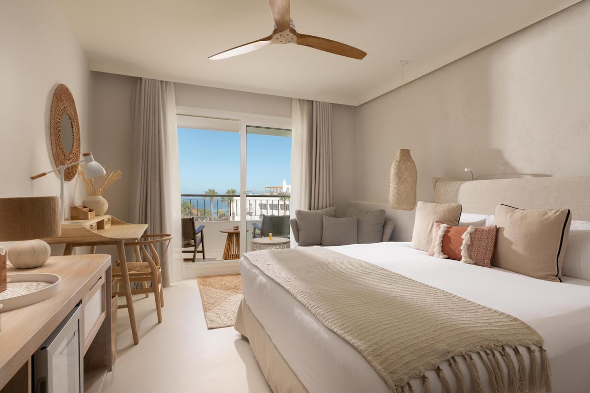 A hotel room with a large bed, a desk with a lamp, a chair, and a view of the outdoors.