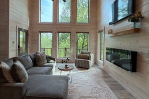 A spacious living room with a grey sectional sofa, a small round coffee table, and a fireplace. Large windows provide a view of the surrounding trees. - 2BR Modern Retreat with 2 Ensuite Baths (Broken Bow)