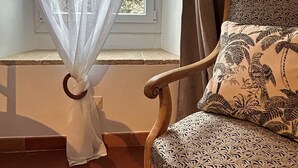 A room with a window covered by sheer curtains, a wooden chair with a patterned cushion, and a tiled floor.