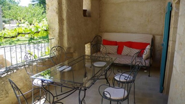 A balcony with a glass table and chairs, a sofa with red cushions, and a view of greenery.
