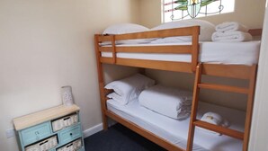 Ein Stockbett mit weißer Bettwäsche und Handtüchern, eine kleine, blaue Kommode mit Körben und ein Fenster mit dekorativer Gestaltung.