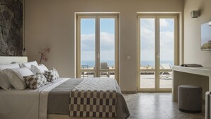 A bedroom with a bed, a stone wall, a view of the beach, and a desk with a chair. - Almarossa Villas (Santorini)
