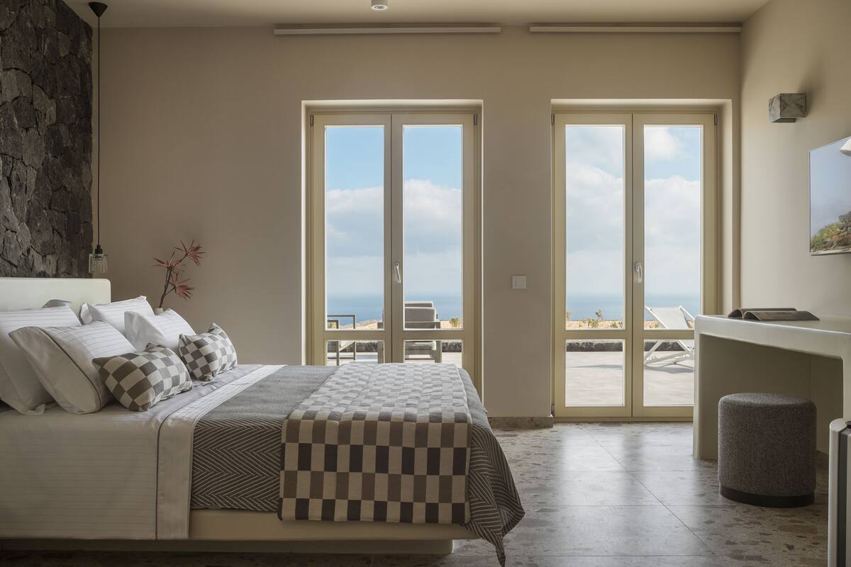 A bedroom with a bed, a stone wall, a view of the beach, and a desk with a chair.