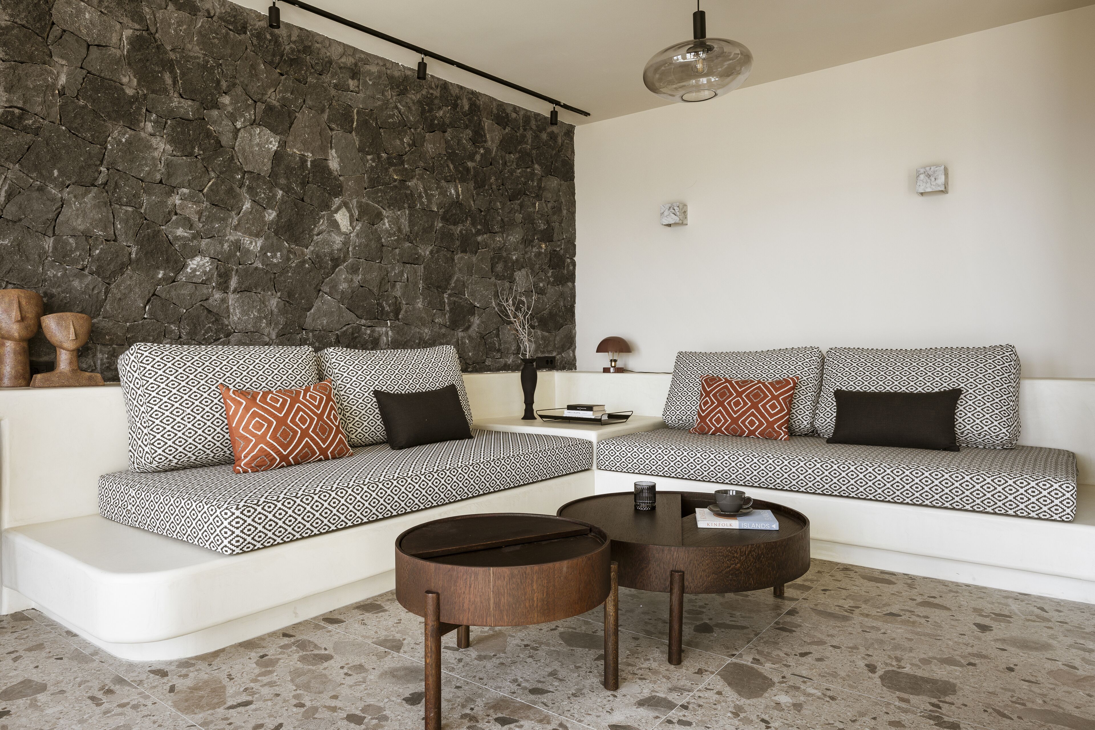 Una sala de estar moderna con una pared de acento de piedra, un sofá blanco con cojines estampados y dos mesas de centro redondas de madera.