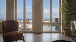 A room with a large sliding glass door leading to a balcony with a view of the ocean. There is a chair and a potted plant inside the room. - Almarossa Villas (Santorini)