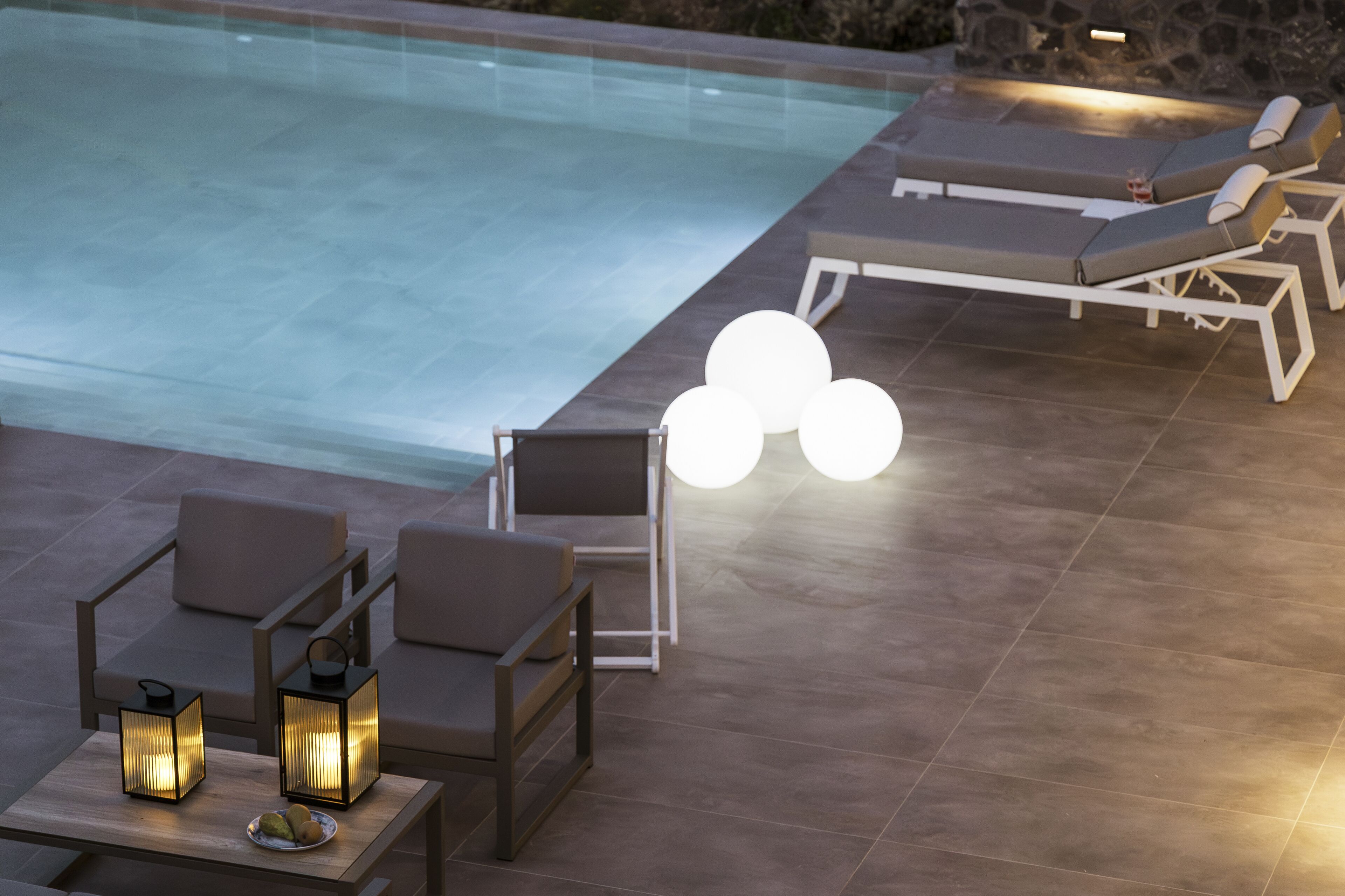 A poolside area with lounge chairs, a table with lanterns, and a tiled floor.