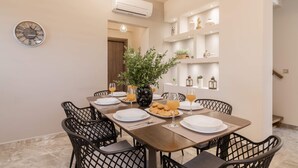 A dining area with a long table, black chairs, and a potted plant. There is a clock on the wall and shelves with decorative items. - Lazai Villa (Zakynthos)