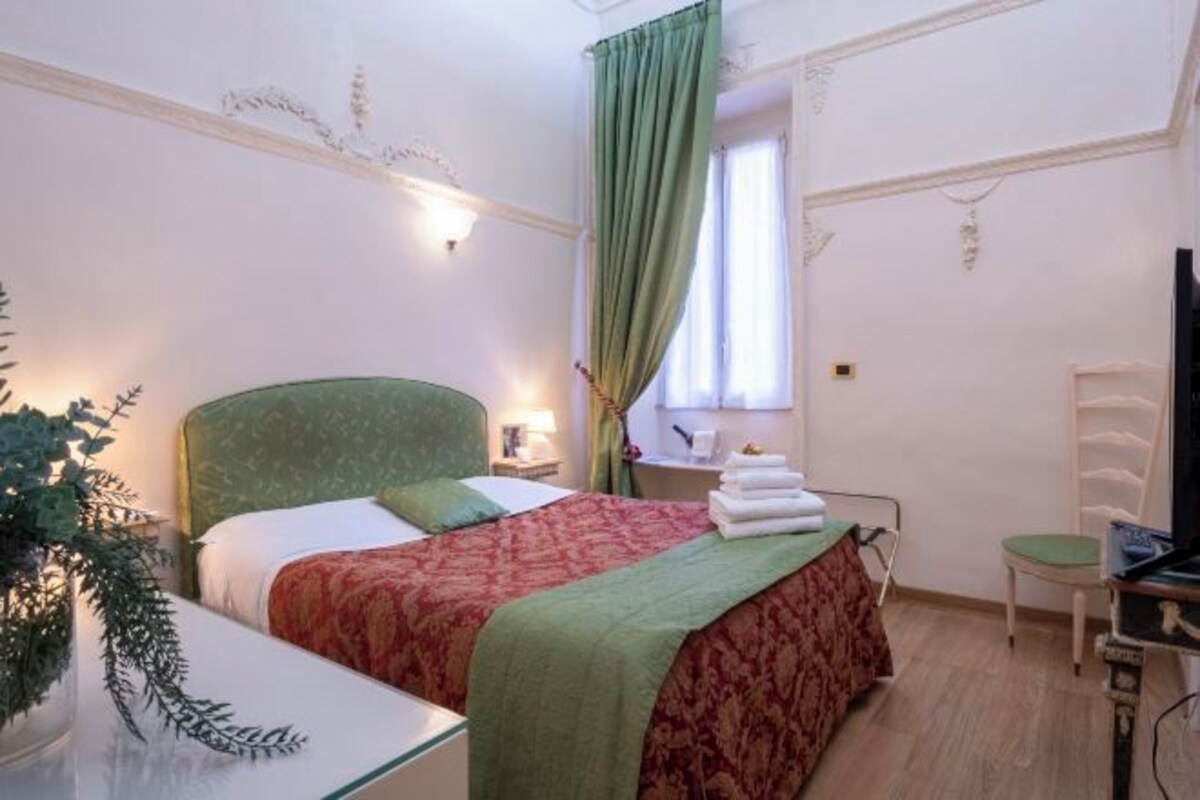 Habitació d'hotel amb un llit, una cadira, un escriptori amb un ordinador i una finestra amb cortines.