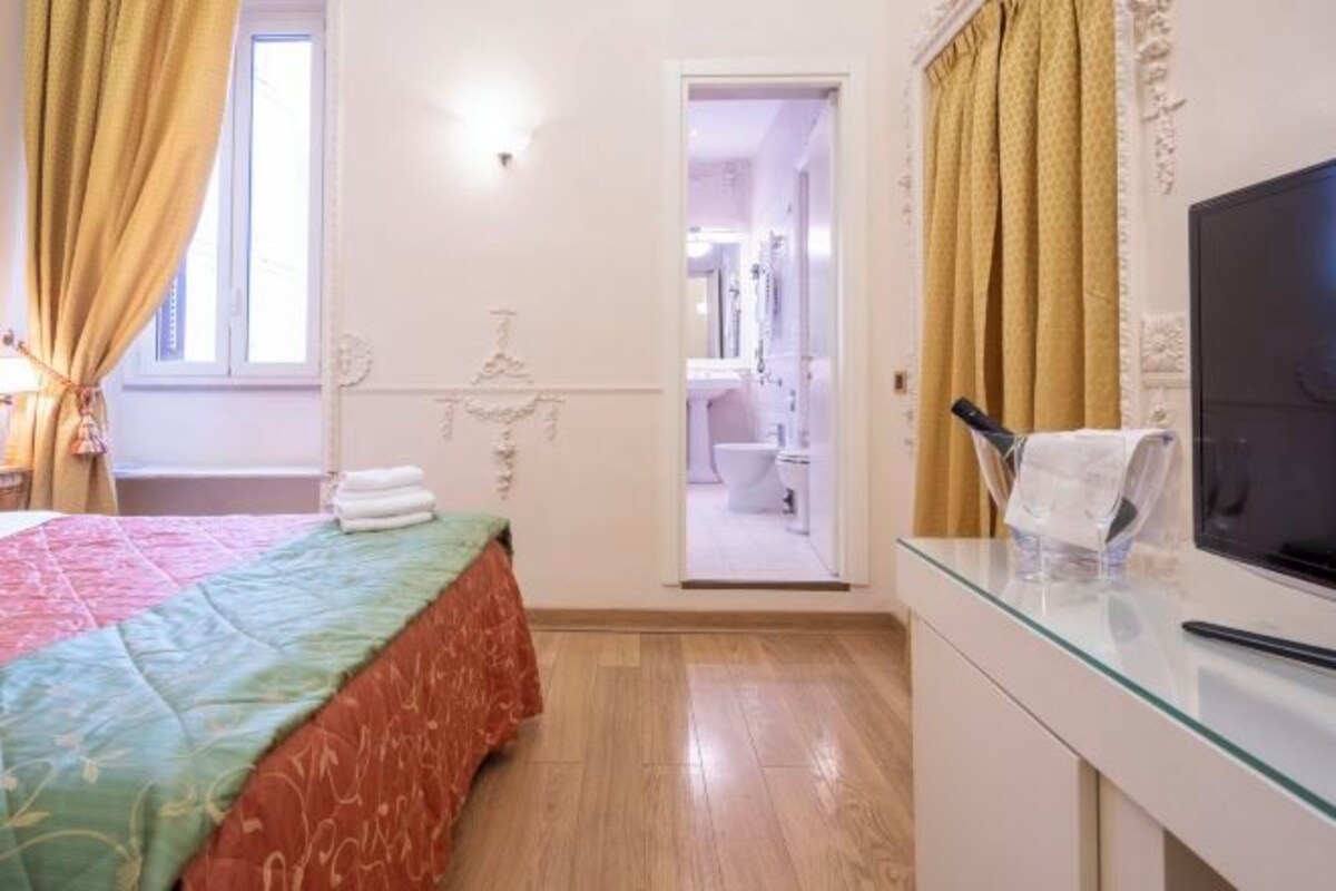 Una habitació d'hotel amb un llit, televisi ó, taula de vidre i un bany visible a través d'una porta oberta.
