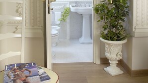 A hallway leading to a bathroom with a white sink and mirror. There is a potted plant on the right and a magazine on a chair to the left. - Suites Rome (Rome)