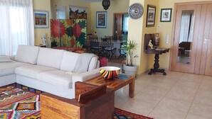 Un salon avec un canapé d’angle blanc, une table basse en bois et un tapis coloré. Au fond, on aperçoit un coin repas avec une table et des chaises.