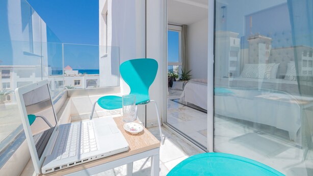 Een balkon met een laptop, turquoise stoelen en een glazen tafel.