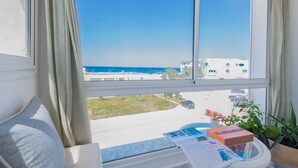 Een kamer met een groot raam met uitzicht op een strand en gebouwen. Er is een vensterbank met een kussen, een kleine tafel met een plantje en een paar voorwerpen op de tafel.