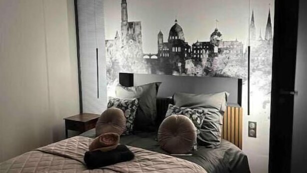 Un dormitorio con cama, mesita de noche, lámpara y un mural de paisaje urbano en la pared.
