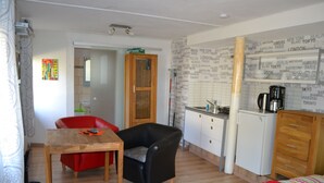 Quarto compacto com mesa de jantar de madeira, uma cadeira de couro preto, sofá vermelho, pequeno espaço de cozinha e uma cama com cobertor vermelho.