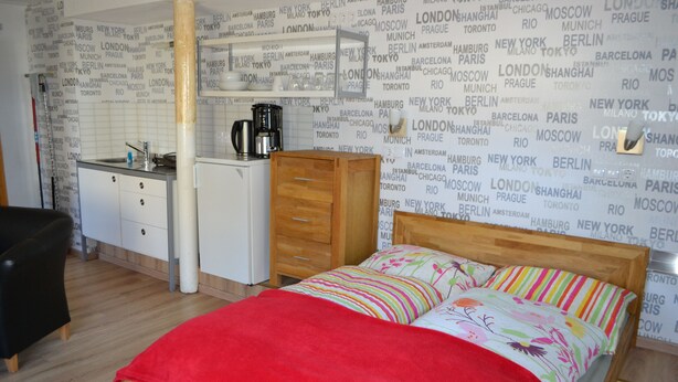 Quarto com cama, cómoda de madeira, pequeno kitchenette e parede com nomes de cidades.