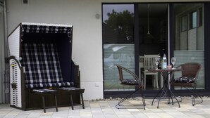 An outdoor seating area with a covered chair and two chairs with cushions, a table with drinks, and a view of a house through a glass door.