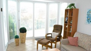 A living room with a sofa, armchair, and ottoman, a wooden bookshelf, and a large window with blinds. - Appartement "ordinger Diek" in St. Peter-ording für 2 Personen (St. Peter-Ording)