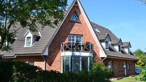A two-story house with a prominent triangular roof, a balcony with a metal railing, and a small window with shutters. - Appartement "ordinger Diek" in St. Peter-ording für 2 Personen (St. Peter-Ording)