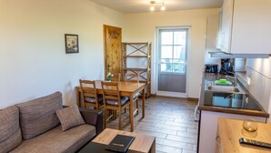 A compact living space with a wooden dining table, four chairs, a sofa, a small kitchen area with a sink and cabinets, and a door leading to an outdoor area. - Appartement "reiterhof Immensee H" in St. Peter-ording für bis zu 5 Personen (St. Peter-Ording)