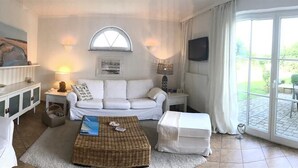 Ein Wohnzimmer mit einem weißen Sofa, einem Couchtisch und einem Esstisch mit Stühlen. Ein Fenster mit Vorhängen, ein Gemälde an der Wand und eine Lampe auf einem Beistelltisch sind ebenfalls vorhanden.