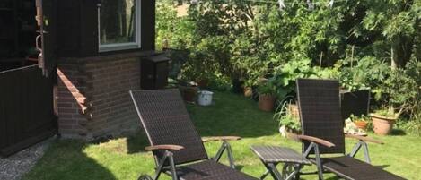 A thatched-roof cottage with a small window and a door, two wicker lounge chairs, and a grassy yard with potted plants.