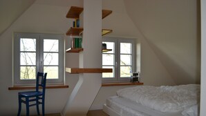 Een slaapkamer met een schuine kap, een bed, een stoel en twee ramen met houten planken erboven.