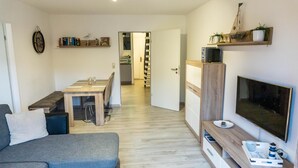 Ein modernes Wohnzimmer mit einer Couch, einem Esstisch und einem Fernseher. Im Hintergrund befindet sich eine Küche mit einer Treppe, die zu einer anderen Ebene führt.