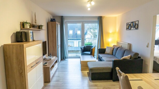 Ein modernes Wohnzimmer mit einer Couch, einem Couchtisch und einem Fernseher auf einem Holzständer. Eine Schiebetür aus Glas führt auf einen Balkon.