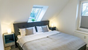 Ein Schlafzimmer mit schrägen Wänden, einem Bett mit gesteppter Bettdecke, zwei Nachttischen mit Lampen und einem Fenster mit Jalousien.
