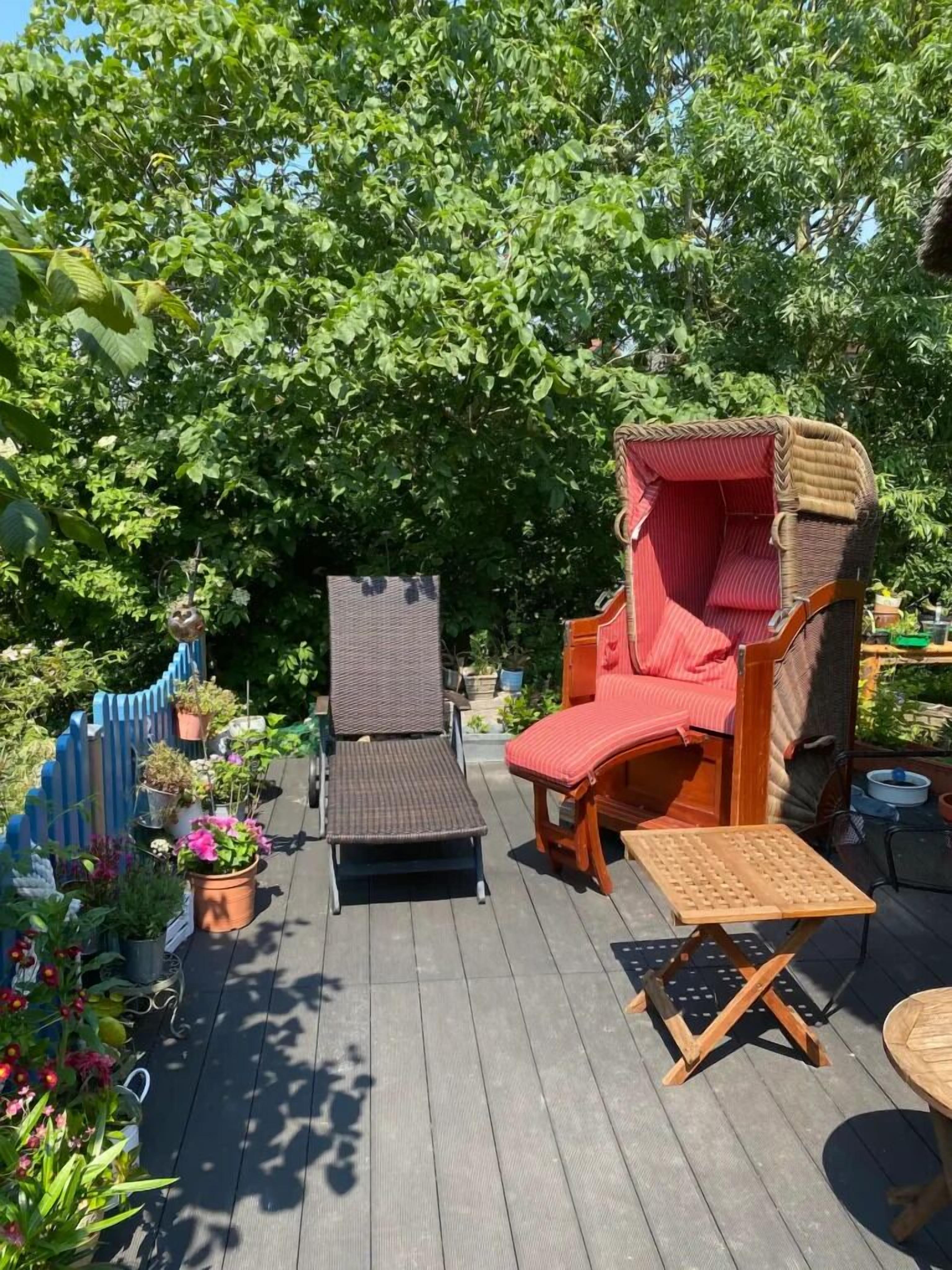 A wooden deck with two lounge chairs, one with a red cushion and the other with a brown cushion. There are potted plants and a blue fence in the background.