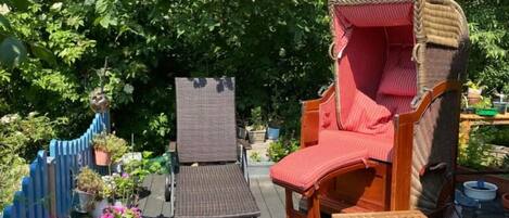 A wooden deck with two lounge chairs, one with a red cushion and the other with a brown cushion. There are potted plants and a blue fence in the background.