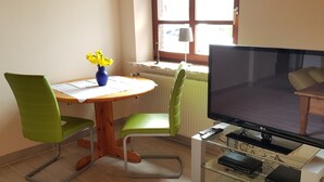 A living room with a wooden table, green chairs, a TV, and a glass cabinet with electronic devices.