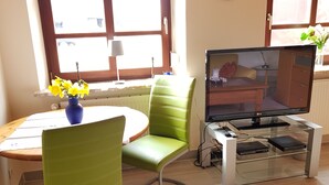 A modern living room with a round dining table, green upholstered chairs, a flat-screen TV on a glass stand, and a vase with yellow flowers.