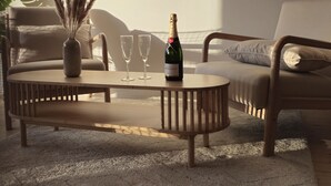 Un salon moderne avec une table basse ronde en bois, une bouteille de champagne et deux verres. Un grand miroir orne le mur et une plante se trouve dans un coin.