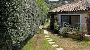 Un sendero conduce a una casa de techo con tejas, rodeada de setos cuidados y un pequeño jardín con flores.