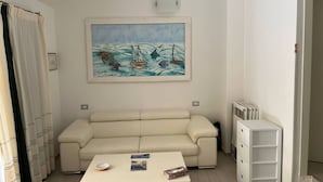 Una habitación con un sofá color beige, una mesita con un cuenco y revistas, un cuadro de barcos sobre fondo azul y un radiador blanco.